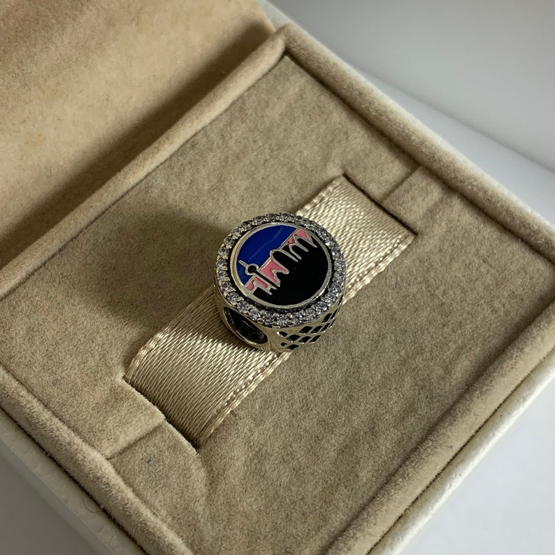 Puede incluir: Un charm de plata con un dise&ntilde;o de ciudad en esmalte azul y rosa. El charm est&aacute; rodeado de piedras de circonita c&uacute;bica transparentes.