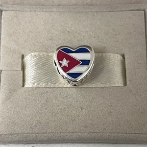 Puede incluir: Un charm de plata en forma de corazón con un diseño de esmalte azul, blanco y rojo que representa la bandera cubana. El charm presenta una estrella blanca sobre un triángulo rojo.