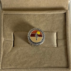 以下が含まれることがあります： 砂漠の風景とサボテン、沈む太陽をカラフルなエナメルで描いたシルバー製のチャーム。チャームは透明なキュービックジルコニアの石で囲まれています。