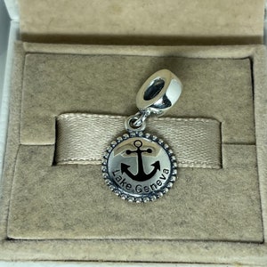Puede incluir: Un charm de plata con un ancla negra y el texto "Lake Geneva" sobre un fondo blanco. El charm est&aacute; en una peque&ntilde;a caja.