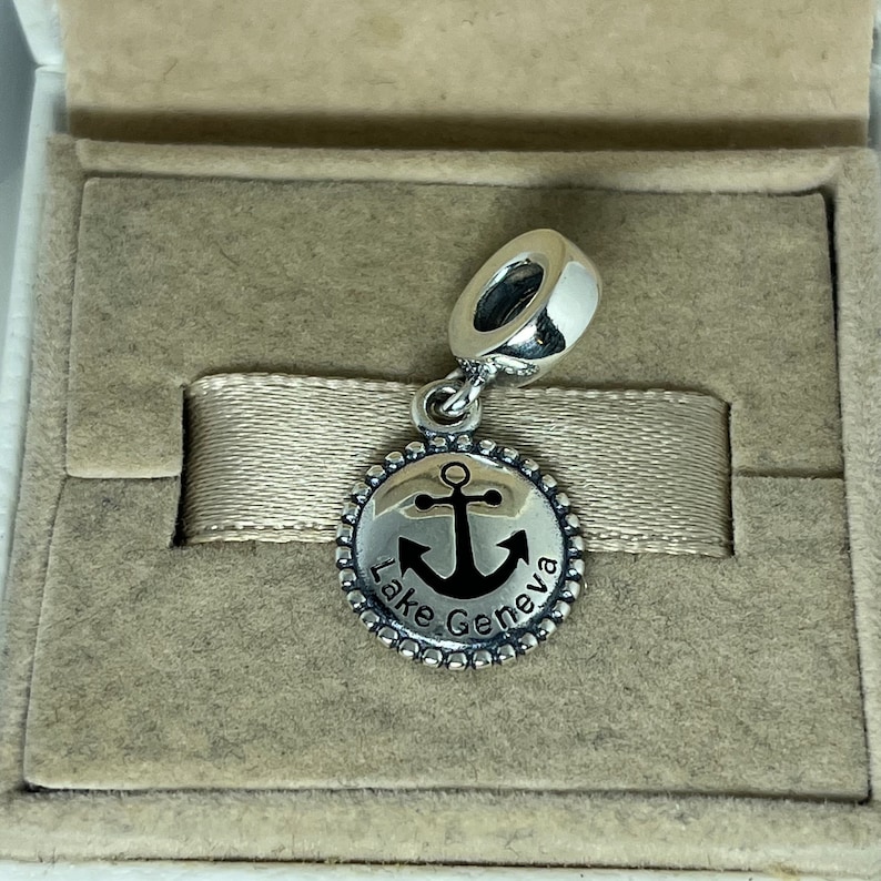 Puede incluir: Un charm de plata con un ancla negra y el texto "Lake Geneva" sobre un fondo blanco. El charm est&aacute; en una peque&ntilde;a caja.