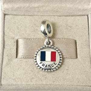May include: Silver charm featuring a round, white, blue and red enamel French flag design. The charm has a beaded border and the word "FRANCE" written below the flag.