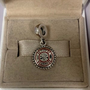 May include: Silver charm with a red and black enamel fire department logo. The logo features a fireman's helmet, ladder, hose, and axe. The charm is in a white box with a white ribbon.