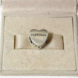 May include: A silver heart-shaped charm with the word "PORTUGAL" engraved on the front. The charm has a decorative beaded edge and is presented in a beige box with a ribbon.