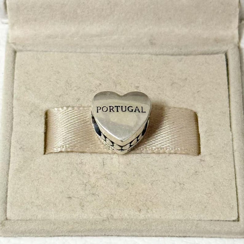 May include: A silver heart-shaped charm with the word "PORTUGAL" engraved on the front. The charm has a decorative beaded edge and is presented in a beige box with a ribbon.