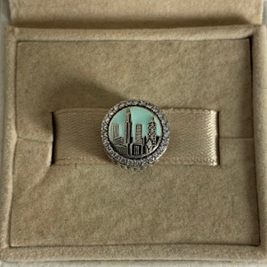 May include: A silver charm with a blue and white enamel cityscape design, surrounded by a clear crystal halo.