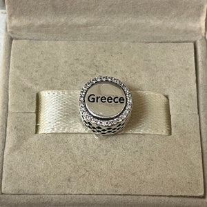 May include: A silver charm with a circle design and clear crystals around the edge. The word "Greece" is printed in black inside the circle.