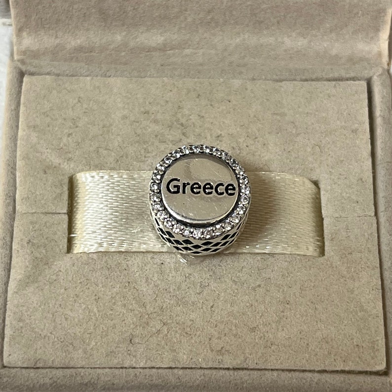 May include: A silver charm with a circle design and clear crystals around the edge. The word "Greece" is printed in black inside the circle.
