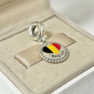 Pode incluir: Berloque de prata com um design de esmalte em forma de cora&ccedil;&atilde;o que apresenta as cores da bandeira belga: preto, amarelo e vermelho. O berloque tem uma borda de contas e a palavra "B&eacute;lgica" inscrita abaixo do cora&ccedil;&atilde;o.