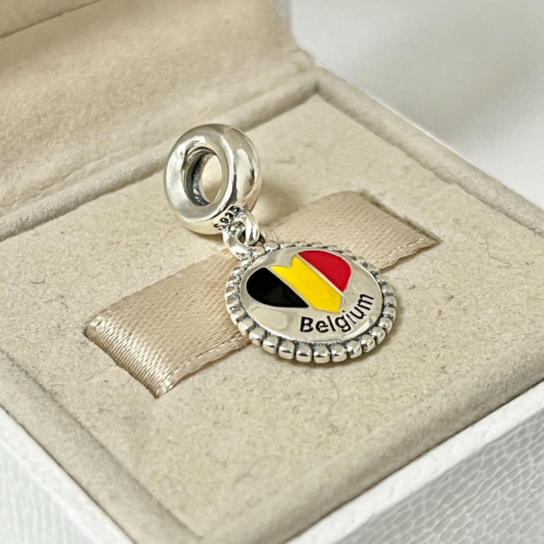 Pode incluir: Berloque de prata com um design de esmalte em forma de cora&ccedil;&atilde;o que apresenta as cores da bandeira belga: preto, amarelo e vermelho. O berloque tem uma borda de contas e a palavra "B&eacute;lgica" inscrita abaixo do cora&ccedil;&atilde;o.