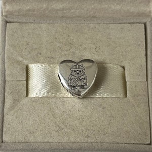 May include: A silver heart-shaped charm featuring a detailed engraving of a cartoon character wearing a hat and holding a cookie.