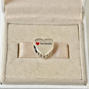 May include: Silver charm in the shape of a heart with a red heart and the text "Bermuda" on it.