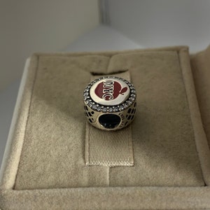 May include: A silver charm featuring a red apple with the text "NYC" in white. The charm is adorned with clear cubic zirconia stones.