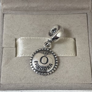 May include: Silver charm with a round, textured disc featuring the word "Pandora" and a crown above the letter "O".