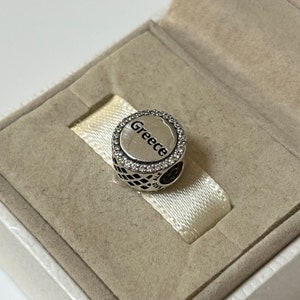 May include: Silver charm with a white and black checkered pattern and a circle with the word "Greece" in black lettering surrounded by clear crystals.