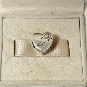 Könnte beinhalten: Ein silberner herzförmiger Charm mit einer schwarzen Umrisslinie von Marokko und einem roten Herz. Das Wort "Morocco" ist in Schwarz geschrieben. Der Charm befindet sich in einer hellbeigen Box mit einem beigen Band.