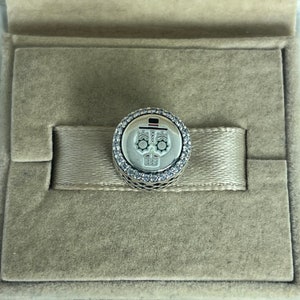 May include: A silver charm featuring a Day of the Dead sugar skull design. The round charm has a white skull design in the centre, encircled by a ring of clear crystals. The charm is displayed in a beige jewellery box.