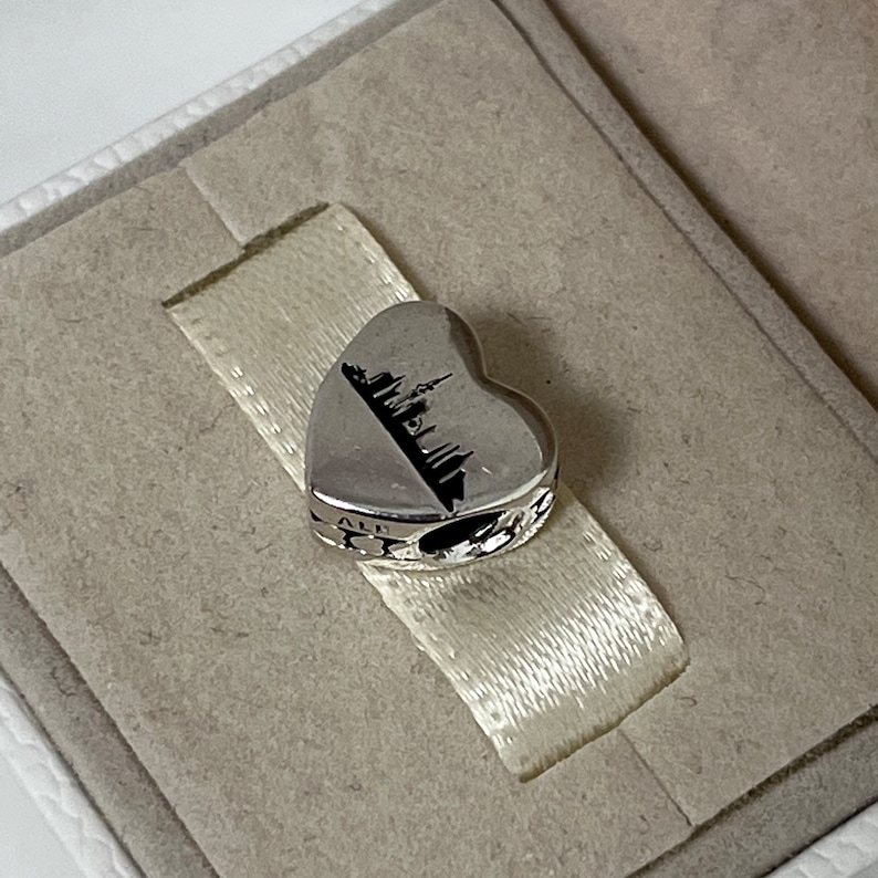 Może przedstawiać: Srebrny charm w kształcie serca z wygrawerowaną sylwetką miasta na przodzie. Charm ma mały otw&oacute;r u g&oacute;ry do przymocowania do bransoletki.