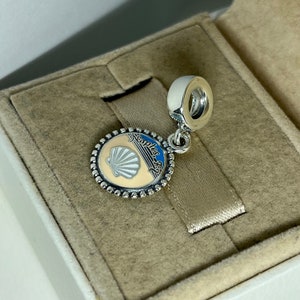 May include: Silver charm with a white seashell and blue and white stripes on a beige background. The charm has the text "Naples, FL" around the edge.