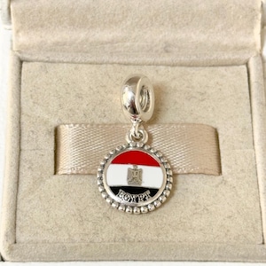 May include: Silver charm with a round enamel pendant featuring the flag of Egypt. The pendant has a red, white, and black enamel design with a gold eagle emblem and the word "EGYPT" in black lettering.