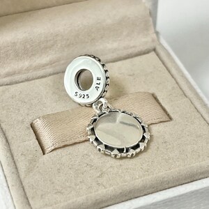 May include: A silver charm with a round, open-faced design and a decorative border. The charm is marked with "S925 ALE" on the back.