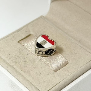 Puede incluir: Charm en forma de coraz&oacute;n con el dise&ntilde;o de la bandera egipcia en rojo, blanco, negro y plateado. El charm tiene la palabra "ALE" en el lateral y se presenta en una caja de joyer&iacute;a.