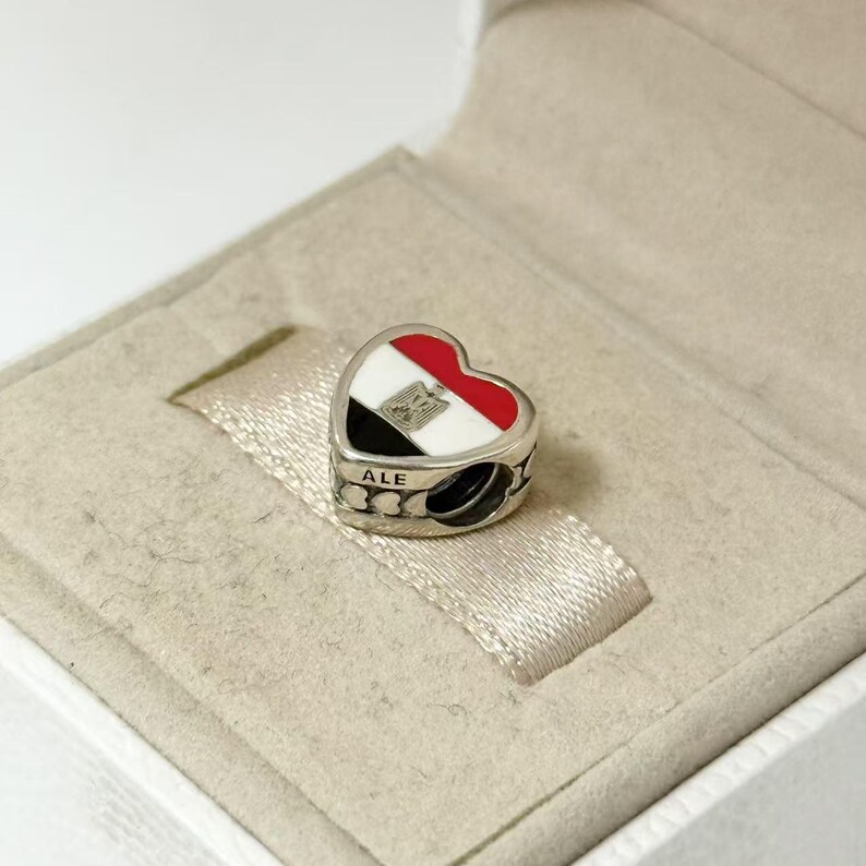 Puede incluir: Charm en forma de coraz&oacute;n con el dise&ntilde;o de la bandera egipcia en rojo, blanco, negro y plateado. El charm tiene la palabra "ALE" en el lateral y se presenta en una caja de joyer&iacute;a.