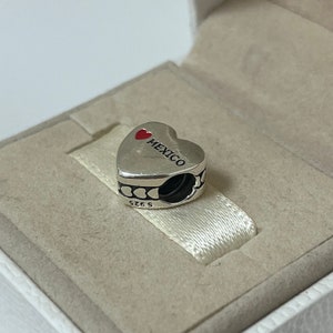 Puede incluir: Encanto de plata en forma de coraz&oacute;n con un coraz&oacute;n rojo y la palabra "MEXICO" en &eacute;l. El encanto tiene un peque&ntilde;o lazo de plata en el lateral.