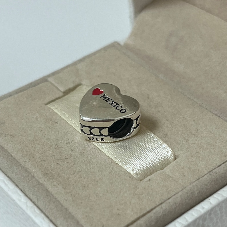 Puede incluir: Encanto de plata en forma de coraz&oacute;n con un coraz&oacute;n rojo y la palabra "MEXICO" en &eacute;l. El encanto tiene un peque&ntilde;o lazo de plata en el lateral.