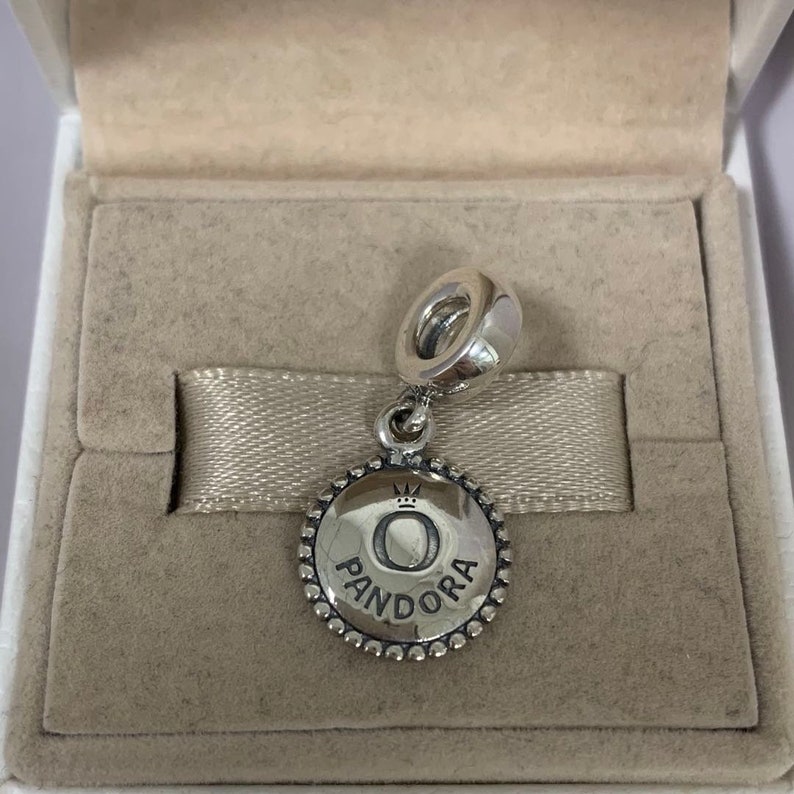 Puede incluir: Un charm de plata con un disco redondo de plata que muestra la palabra "Pandora" y una corona sobre la letra "O".