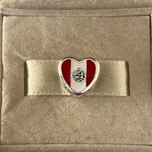 May include: A silver heart charm with red and white enamel, depicting the flag of Peru. The charm features the national coat of arms in the center.