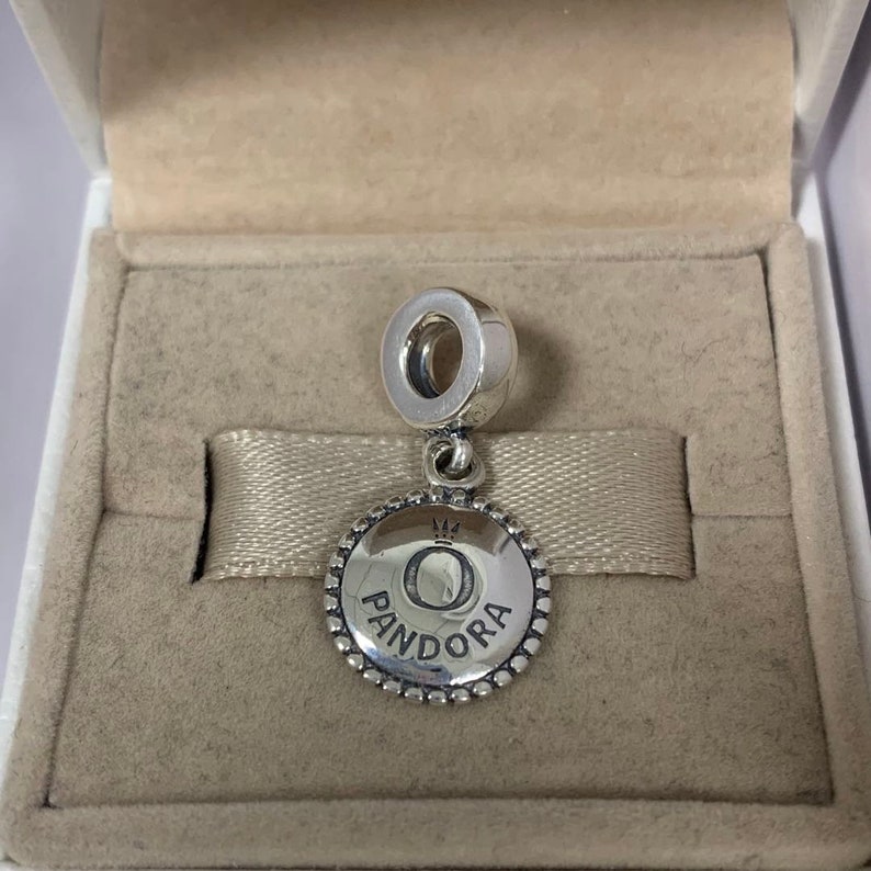 May include: A silver Pandora charm with a circular pendant displaying the letter "O" and the brand name "Pandora". The charm hangs from a loop and is presented in a beige jewellery box.