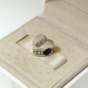 Puede incluir: Un charm plateado en forma de coraz&oacute;n con un dise&ntilde;o de sirena. El charm est&aacute; en una caja blanca con una cinta de sat&eacute;n beige. El charm tiene la palabra "ALE" grabada.