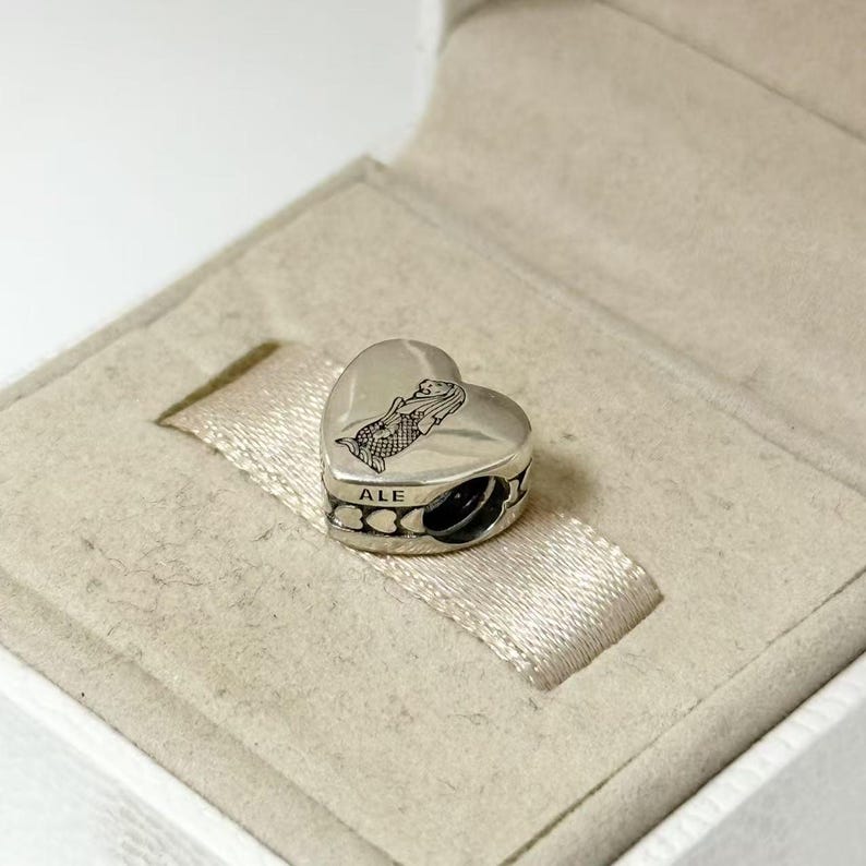 Puede incluir: Un charm plateado en forma de coraz&oacute;n con un dise&ntilde;o de sirena. El charm est&aacute; en una caja blanca con una cinta de sat&eacute;n beige. El charm tiene la palabra "ALE" grabada.