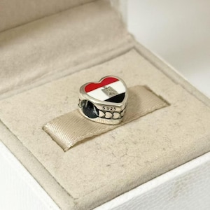 Puede incluir: Un charm en forma de coraz&oacute;n con el dise&ntilde;o de la bandera egipcia. El charm presenta un esquema de color rojo, blanco y negro con el escudo de armas egipcio. El charm es de plata y est&aacute; marcado con "S925". El charm se presenta en una caja blanca.