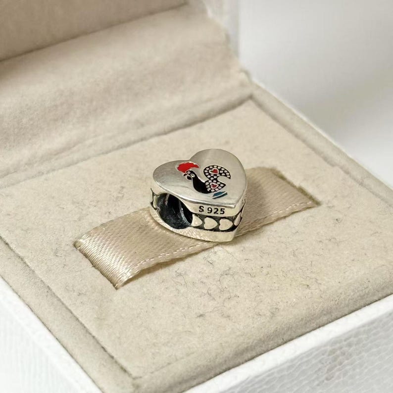 May include: A heart-shaped silver charm with a rooster design in red, black, and white. The charm has a small loop for attaching to a bracelet or necklace. The charm is in a white box with a beige ribbon.