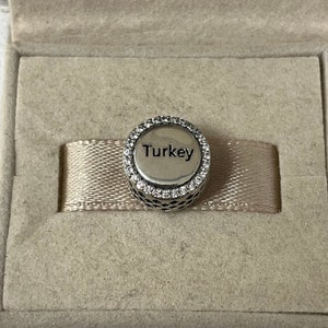 May include: A silver charm with a circle of clear stones around the edge. The charm has the word "Turkey" written on it.