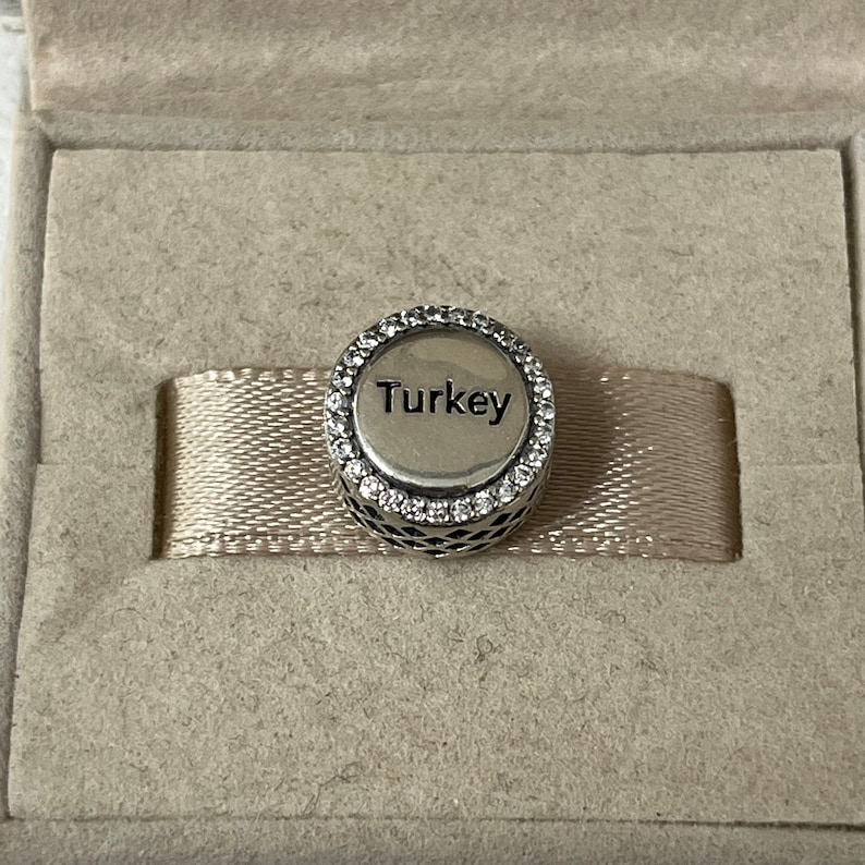 May include: A silver charm with a circle of clear stones around the edge. The charm has the word "Turkey" written on it.