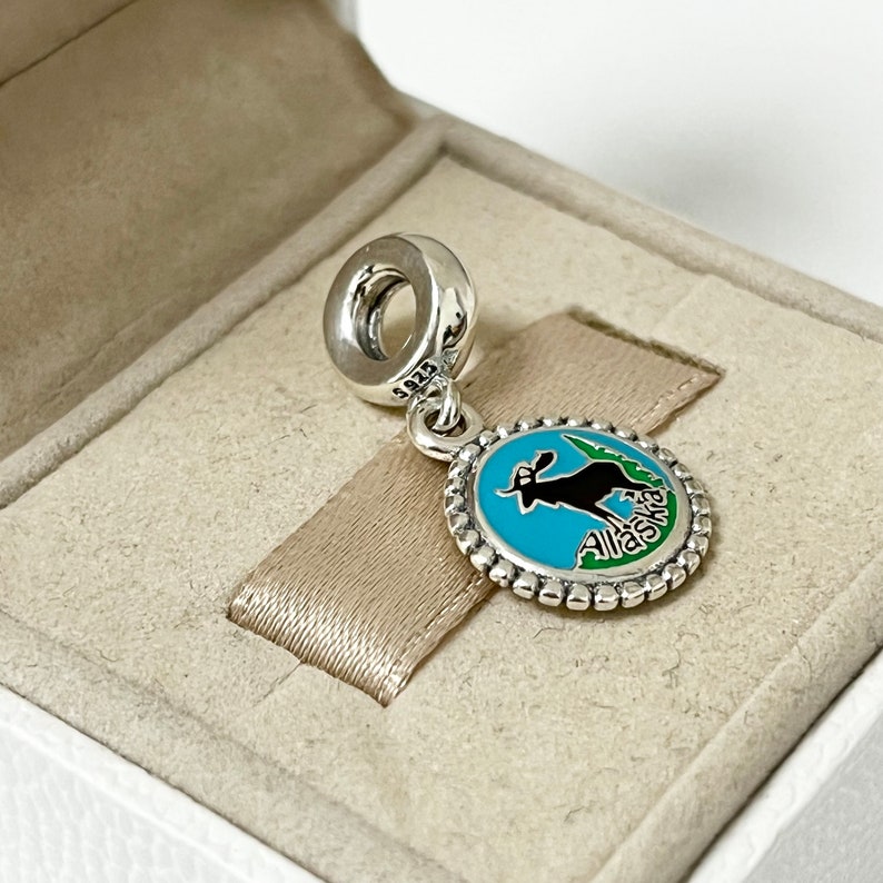 May include: Silver charm with a blue and green enamel design featuring a silhouette of a moose and the word "Alaska".