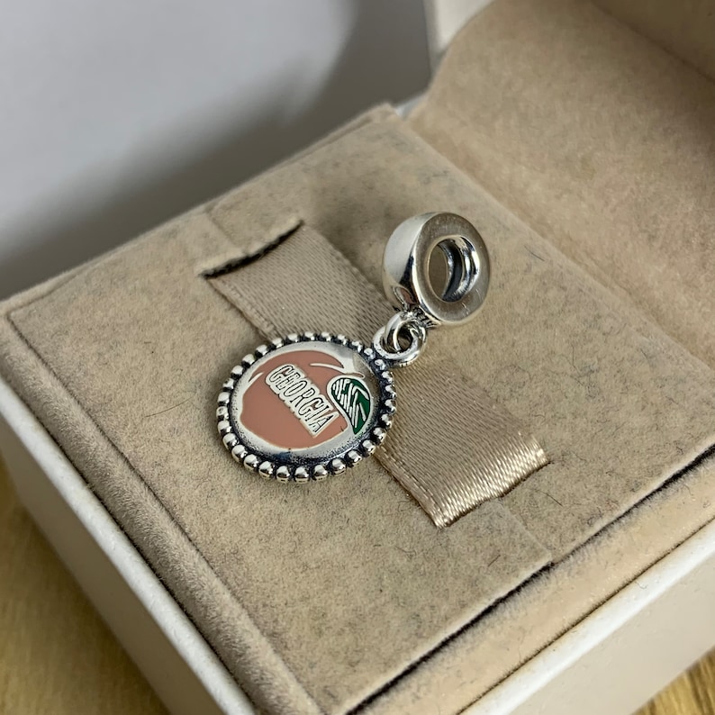 May include: Silver charm with a peach design. The peach is pink and has the word "Georgia" written on it. The charm has a green leaf and a silver bead border.
