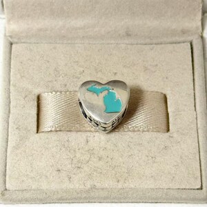 Peut inclure: Un charm argenté en forme de cœur avec le contour turquoise de l'État du Michigan. Le charm est présenté dans un écrin beige avec un ruban. Le charm porte les mots "ALL" et "925" sur le côté.