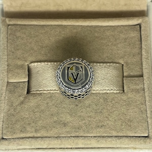 May include: A silver charm with a black and gold Vegas Golden Knights logo surrounded by clear rhinestones.