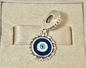 May include: A silver charm with a blue and white evil eye design. The charm is in a small box with a light brown ribbon.