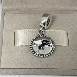 Peut inclure: Charme en argent avec un logo de lion noir sur un disque rond avec une bordure perlée. Le charme est conçu pour être ajouté à un bracelet ou un collier.