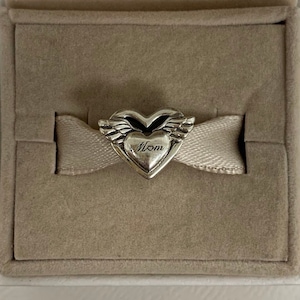 May include: A silver heart-shaped charm with the word "Mom" engraved on it. The charm features small wings on either side of the heart and is displayed on a light beige ribbon inside a gray box.
