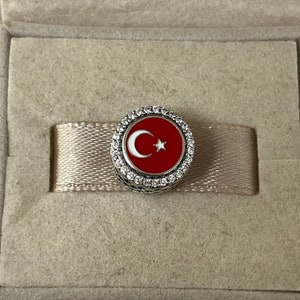May include: A silver charm featuring a red enamel Turkish flag design. The flag depicts a white crescent moon and a white star. The charm is encircled by a ring of clear cubic zirconia stones.
