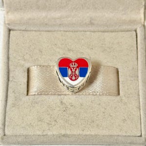 Puede incluir: Un charm en forma de corazón con el diseño de la bandera serbia, con esmalte rojo, azul y blanco. El charm está hecho de metal plateado y se presenta en una caja de joyería beige.