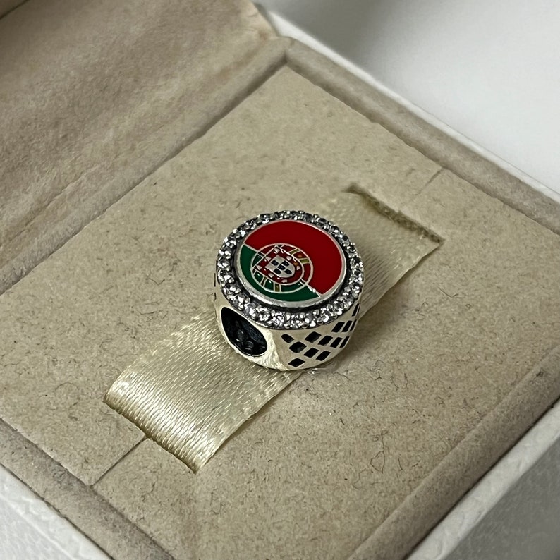 Puede incluir: Un charm de plata con un dise&ntilde;o de esmalte rojo, verde y blanco de la bandera portuguesa. El charm est&aacute; rodeado por un c&iacute;rculo de piedras de circonita c&uacute;bica transparentes.