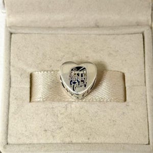May include: A heart-shaped silver charm featuring a detailed illustration of a tuk-tuk, a three-wheeled auto rickshaw. The charm is set against a neutral-toned box, highlighting the intricate design and craftsmanship of the jewelry.