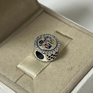 May include: A silver charm with a round, black and white patterned base and a circular top featuring a colorful illustration of two people in traditional Japanese clothing. The charm is surrounded by a ring of clear cubic zirconia stones.